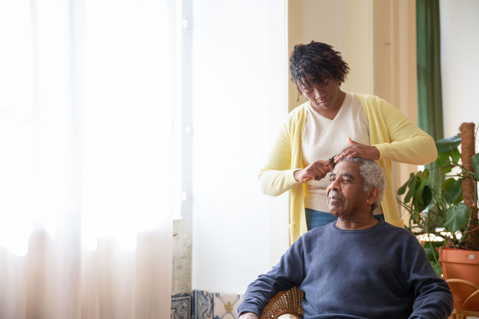 A caregiver gently grooming an elderly man beside a sunlit window